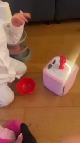 A baby is sitting on a wooden floor, near a pink square object with a red figure on top. The baby is wearing white pajamas with thin gray stripes. To the left, there's a red bowl and a toy ball run with white and green pieces.  The baby reaches for the red figure on the pink object and touches it. There is a stuffed animal at the bottom left of the frame. The background shows a dark blue couch.