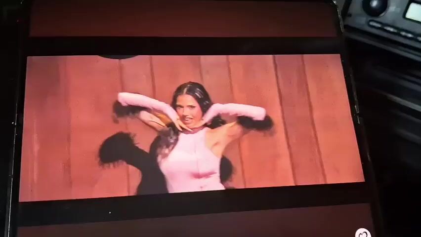 The video displays a screen showing a performance. A woman in a pink top and gloves is dancing energetically. She has long, dark hair and tattoos on her arms. Behind her, another person with blonde hair and dark makeup can be seen. The background appears to be a room with wood-paneled walls and a clock on the wall. The lighting is warm and theatrical.