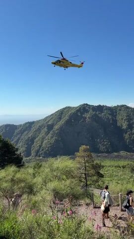 A yellow helicopter flies over a mountainous, wooded area under a clear blue sky. In the foreground, a dirt path is visible, with several people walking on it. One person in the foreground is wearing a backpack and appears to be looking towards the helicopter. There are also plants and flowers along the path. The background shows rolling green hills and a distant hazy landscape.