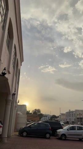 The video shows a daytime street scene with several cars parked along the curb. A light-colored building with arched windows and columns is on the left side of the frame. The sun is setting in the distance, partially obscured by a tree, casting a golden glow on the scene. The sky is filled with light, fluffy clouds. Streetlights are visible along the street, and there are other buildings on the opposite side.