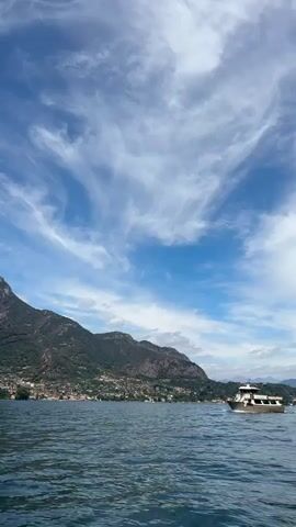 A wide shot captures a boat moving across a body of water. The boat is light brown with white accents and appears to have multiple decks. In the background, a mountain range is visible, covered in trees and dotted with buildings. The sky is blue with wispy white clouds. The water is a deep blue with gentle ripples. The perspective is low, suggesting the footage was taken from another boat or the water's surface.