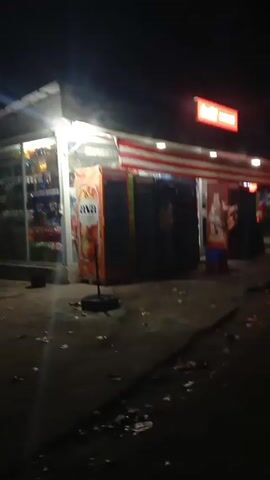 The video begins with a dark, shaky shot of a convenience store at night. The store has a red awning with white stripes, and a neon sign above that reads "Daily Market". Several soda fridges are visible in front of the store, one displaying an "Ava Lemonade" advertisement. The ground is littered with trash. The camera then moves away from the store and towards a dark street with some distant lights. As the camera moves further down the street, the scene becomes increasingly blurry and dark, with only the faint lights of buildings and traffic signals visible in the distance.