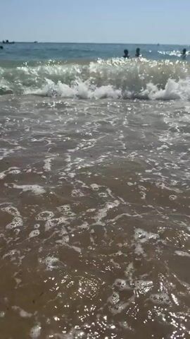 The video captures a low-angle perspective of gentle waves rolling onto a sandy beach. The water is a clear, slightly murky brown, and the sand is a darker brown, appearing wet and packed near the shoreline. As the waves approach, they form white crests of foam, some of which break and dissipate, creating ripples and small bubbles on the surface. In the background, the ocean extends to the horizon, with a clear, pale blue sky above. Several dark silhouettes of people can be seen in the water in the distance, suggesting they are swimming or wading. The overall scene depicts a serene and inviting beach environment.