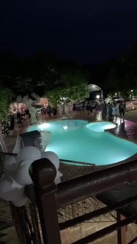 This video shows a nighttime outdoor party scene with a swimming pool as the central focus. Several people, dressed in formal attire, are gathered around the pool, some are standing in groups and interacting. The pool is illuminated with a bright turquoise light, and the surrounding area is also lit by several artificial lights, casting shadows on the wet ground, suggesting it may have recently rained or the area has been recently watered. Trees and foliage form a dark backdrop against the night sky, and a fence or railing with a brown wooden top is visible in the foreground, obscuring the lower part of the scene. White balloons are clustered near the railing. The overall atmosphere appears festive.