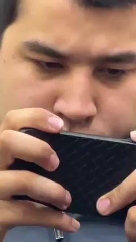 A close-up shot shows a person intensely focused on a smartphone screen. The person holds the phone horizontally with both hands, thumbs actively engaged on the screen. The phone has a black, textured case. The individual has dark hair, dark eyebrows, and is wearing a gray shirt or jacket with a visible zipper pull. The background is out of focus.