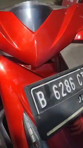 This media file features a close-up view of a red and black scooter. The scooter is the primary object of focus, with its shiny red bodywork and black seat with red accents clearly visible. The license plate of the scooter is visible, displaying the text "B 6286 CT". The scooter is parked on a concrete surface, and the background is dimly lit, suggesting it might be nighttime or indoors. There are some indistinct objects in the background, including what appears to be a red box with some lettering on it, possibly from a store or delivery service, and some pipes. The overall scene is static, with no people or apparent activity taking place. The focus is entirely on the details of the scooter, highlighting its color and design. The lighting creates a slight sheen on the scooter's surface, emphasizing its polished appearance.