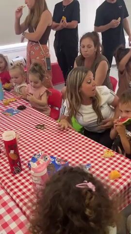 The video shows a group of young children and adults gathered around a table covered with a red and white checkered tablecloth. The children are seated and appear to be eating snacks, with some holding pieces of fruit and cookies. One child is eating watermelon. Several adults are standing behind the children, also holding snacks, and seem to be supervising the event. The setting appears to be indoors, possibly a community hall or a decorated room, with white walls and some colorful plastic chairs. The lighting suggests it could be daytime.

Notable details include:
- The children are of varying young ages, with some appearing to be toddlers.
- Snacks on the table include waffles, cookies (possibly Oreos, as a box is visible), and watermelon.
- A pink water bottle with a "Hello Kitty" design is on the table.
- A red tube-shaped container, possibly for snacks or drinks, is also visible.
- The adults are casually dressed.
- The overall atmosphere seems informal and cheerful, consistent with a children's party or gathering.

Visible text:
- "OREO" is visible on a blue box of cookies.
- A red and white tube has some text that is difficult to read but appears to be in English and possibly Hebrew.
- A pink water bottle has "Hello Kitty" branding.