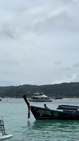 A longtail boat with a person sitting in it floats on the water. In the background, another boat is visible, as well as a lush green island with buildings. The sky is cloudy.