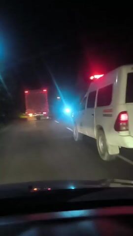 A white van, possibly an ambulance due to a red flashing light on its roof, is driving down a dark road at night, following a large red and white truck. The truck has its brake lights and hazard lights illuminated. The camera is positioned inside another vehicle, looking out through the windshield. The road appears to be paved and is marked with white lines. Trees are visible on the left side of the road. The environment is dark and rural.