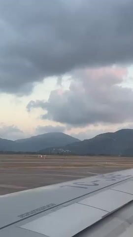A static low-angle shot from the perspective of an airplane window facing the wing shows a runway and buildings. The airplane wing is white with blue text. Beyond the runway are hills covered in buildings and trees. The sky is cloudy with some pink highlights. Another plane with a red tail is visible in the distance on the runway.