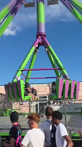 A group of young people are standing in the foreground, looking at a tall amusement park ride. The ride, with its purple and green structure, is in motion. Several people are seated on the ride, their bodies positioned to face outwards. The sky is clear and blue with a few scattered clouds. The ride appears to be at a fair or carnival, with a building visible in the background.