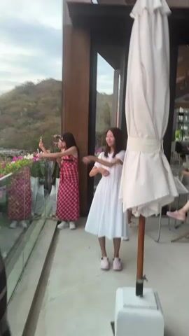 A group of people are standing on a balcony with a scenic view. On the left, a person wearing a red and white checkered dress is holding a phone up to take a picture or video. To the right of that person, a young girl in a white dress is dancing or posing. In the foreground, a large white umbrella is partially closed and tied. The background shows a hillside covered in trees and vegetation. The sky is overcast.