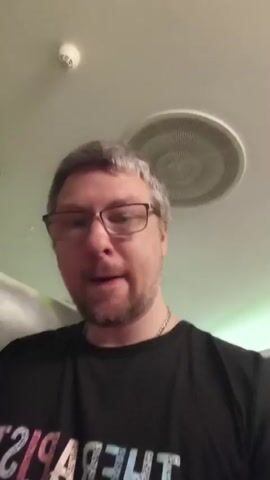 The video shows a medium shot of a man with short, grey hair and glasses, wearing a black t-shirt. He is smiling and looking at the camera. The t-shirt has text on it that reads "THERAPIST Life" in a colorful font, with the word "THERAPIST" appearing in reverse. The background appears to be an indoor setting with a textured wall featuring abstract green and grey designs. A white cylindrical object, possibly a light fixture or pillar, is visible behind the man's right shoulder. The ceiling has several circular light fixtures and a smoke detector. The lighting suggests it is daytime. The man's expression and demeanor convey a sense of warmth and approachability. The overall scene is casual and personal, likely a selfie or a video message. The location is identified as Troisdorf, Germany, but no specific landmarks or contextual clues within the video directly confirm this.