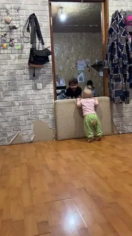 A toddler stands facing away from the camera in front of a doorway, holding onto a tan cushion that is propped up to act as a barrier. The toddler is wearing a pink short-sleeved shirt and green pants with a small floral pattern. Through the doorway, two figures are visible. One is a young boy with dark hair, bent over with his head near the cushion. The second is sitting farther back in the room. The room beyond the doorway has wood-paneled walls and contains furniture and other items. The room the toddler is standing in has laminate wood flooring and a brick-patterned wall on the left, where a black purse and other items are hanging. The wall has visible damage at the bottom. To the right of the doorway hangs a patterned robe or coat on a hook. The video is handheld, with the camera moving slightly.