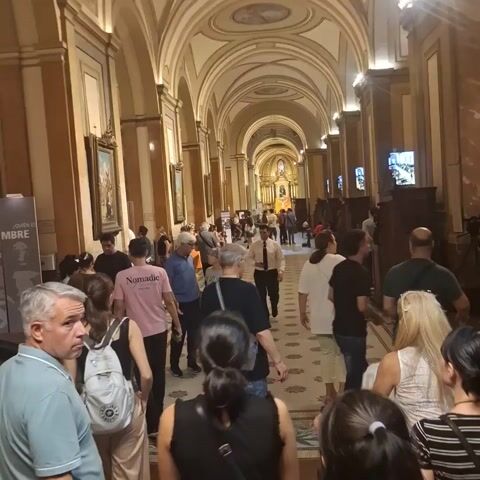 The video shows a crowded hallway with an arched ceiling. Many people are walking down the hallway. There are paintings and screens on the walls. The hallway leads to a bright altar at the end. A few people are seated on the left side of the hallway.