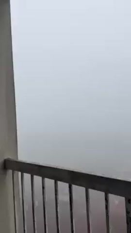The camera is positioned on a balcony, looking out into dense fog that obscures most of the view. In the foreground is the metal railing of the balcony, which is slightly weathered and painted gray. As the camera pans down, glimpses of the ground below become visible, including what seems to be a red surface and some indiscernible figures standing on it. The fog begins to thin slightly as the camera moves to the right, revealing a corner of a white building with flat green rooftops in the distance. The overall atmosphere of the scene is one of limited visibility and stillness due to the heavy fog.