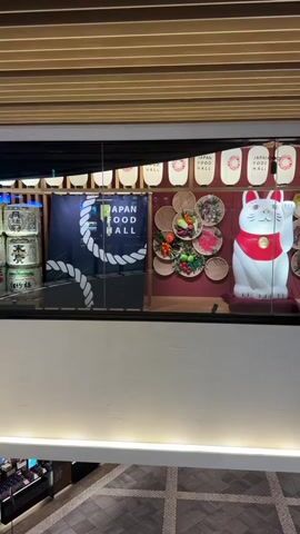 The camera pans across a display for a "Japan Food Hall" located on a higher floor in a shopping center. The display includes a large, illuminated beckoning cat statue, arrangements of baskets filled with food, and a banner that reads "Japan Food Hall" with a rope design. Above the display are several Japanese-style lanterns, each also displaying the words "Japan Food Hall." The space below the display on the lower floor is visible and shows a patterned tile floor and some retail spaces. The upper ceiling is lined with wooden slats. There are no people visible in the scene.