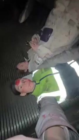 The video shows three people lying on a corrugated surface. The two individuals most visible appear to have blood on their faces. One is wearing a high-visibility jacket over a neon green shirt, and the other wears a white jumpsuit with the number 585 on it. The third person, partially visible, also seems to have blood on their leg. The environment appears dark and possibly industrial.