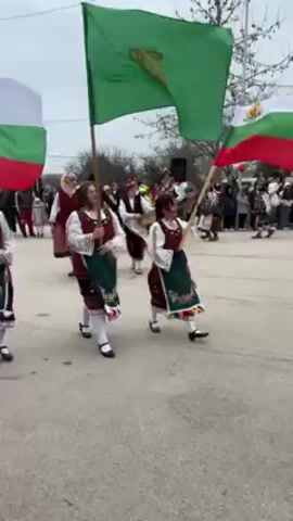 The video shows a group of people in traditional Bulgarian clothing performing a traditional dance. Three women in the foreground are carrying Bulgarian flags. Other participants, including men and women, are wearing vests, skirts, aprons, and flower crowns, and are dancing to music. The setting appears to be a village street, with houses and onlookers in the background. The sky is overcast.