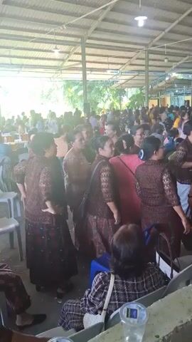This video captures a gathering of people inside a large, open-air structure with a metal roof supported by poles. The space appears to be a community hall or a similar venue. In the foreground, several women are gathered, mostly facing away from the camera and interacting with each other. They are wearing traditional or formal attire, predominantly in shades of brown and maroon, with some individuals in pink. Some of these women are in the process of dancing or moving rhythmically. In the background, a larger crowd of people are visible, milling about and engaging in conversations. The environment suggests a festive or celebratory occasion. Several rows of plastic chairs are visible, some occupied by individuals seated. In the immediate foreground, a woman wearing a checkered top is seated, facing away from the camera. Disposable cups are also visible on the railing in front of her. The lighting appears to be natural, with sunlight filtering in from the sides, indicating it is daytime. There is a general sense of movement and activity throughout the scene.