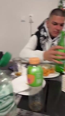 A young man in a black and grey Nike hooded sweatshirt is being filmed holding a water bottle with a green cap, and the label reads "Михалково". He is inside a building with large windows looking out onto an outdoor area with green umbrellas and a sign in Cyrillic. The person filming is holding the water bottle and asks the young man to advertise it. The young man turns and smiles at the camera.