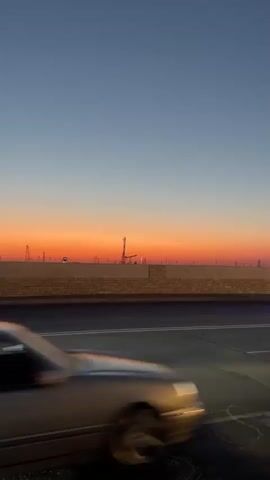 The video shows a highway at sunset. The sky is a gradient of orange and blue. Cars are driving on the highway, their headlights and taillights visible. In the background, there are industrial structures. The camera pans across the scene, capturing the movement of the cars and the changing light.