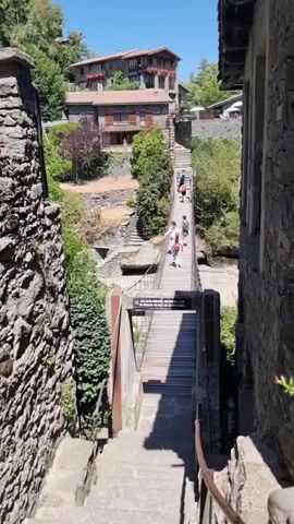 A vertical video shows a stone staircase leading down towards a wooden suspension bridge. Several people, including adults and children, are walking across the bridge. The bridge spans a dry riverbed or ravine. On the other side of the bridge, there are more stone stairs leading up to buildings constructed of stone and wood, with red-tiled roofs. The buildings are nestled on a hillside covered in green trees and vegetation. The sky is clear and blue. The overall scene depicts a historic village or settlement.