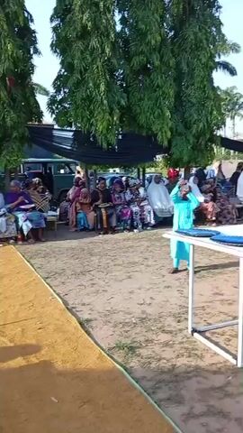 The video shows an outdoor gathering. People are seated in rows on chairs, while others are standing and walking around. There's a table with floral arrangements at the front, and a small child in a blue outfit is walking near it. A rug leads to the chairs, and trees are visible in the background. Some individuals have plates on their laps, and there are tables with cloths on them. The event appears to be taking place during the day with a clear sky.
