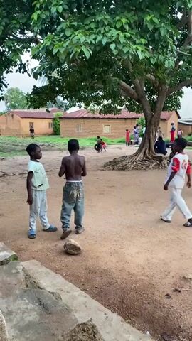 The video captures an outdoor scene in what appears to be a rural village in Riyom, Nigeria. The ground is dirt or sandy earth. In the foreground, three young boys are standing, one shirtless with jeans, another in a light t-shirt and light-colored trousers, and a third in a t-shirt and white trousers. They seem to be interacting playfully or just observing. A small rock is visible on the ground near them.

Dominating the mid-ground on the right is a large tree with expansive, visible roots. Several other children and adults are scattered throughout the background, some sitting beneath the tree's shade, while others walk or stand near the buildings.

The background features several simple, single-story houses constructed from what appears to be mud bricks, topped with corrugated metal roofs. The general atmosphere suggests a casual, everyday village setting. The lighting indicates it is daytime, possibly late morning or afternoon, with diffused light suggesting either an overcast sky or an early/late time of day without direct harsh sunlight. There is no visible text in the frame.