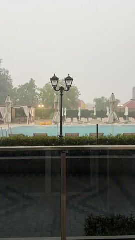 This media depicts an outdoor swimming pool area during a rain shower. The pool is turquoise blue and filled with water, with several lounge chairs and closed umbrellas visible around its perimeter. A black double-headed lamppost stands prominently in the foreground, with a hedge providing a visual barrier. Beyond the pool, trees and buildings can be seen through the rain. The sky is overcast and gray, indicating inclement weather. The scene appears to be a resort or hotel, given the presence of lounge chairs and covered cabanas. The rain is heavy, with visible drops hitting the pool's surface and creating ripples. There is no discernible time of day, but the overcast sky suggests it is not bright daylight. The overall mood is one of a temporarily disrupted outdoor experience due to the weather. No people are visible in the frame. Some text is visible on one of the closed umbrellas, appearing as "VIPERI". The location is Hisarya, Bulgaria.
