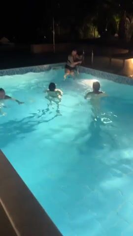 A vertical video shows four women in a swimming pool at night. The pool is illuminated by underwater lights, creating a bright blue glow. Trees and shrubs are visible around the pool, with some outdoor lighting in the background. Three women are swimming or floating in the water. One woman, wearing a dark swimsuit, is standing near the far end of the pool, holding a baby. The woman on the left is swimming on her back, while the other two women in the middle and right are upright in the water. The video briefly captures the sound of water splashing and distant conversation.