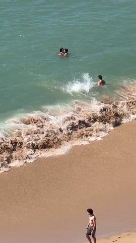 The video shows a beach scene on a sunny day.  The water is light green and waves are breaking on the sandy shore. Several people are in the water: a couple embracing each other is farther out, while a woman swims closer to shore, and a man walks into the water.  Another person is standing on the sand near the bottom of the frame. The camera is positioned at a high angle. The wind noise is noticeable.