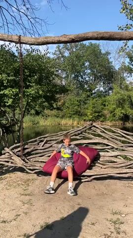 A boy with blonde hair, wearing a grey t-shirt with a yellow design and grey shorts with a white pattern, sits on a large, maroon beanbag. He is positioned in the center of the frame, facing the camera and looking to his right. The beanbag is situated on a sandy patch of ground. Behind him is a lake, bordered by a rustic structure made of intertwined branches. A large, thick tree branch extends horizontally across the top of the frame, with a chain hanging down from it on the left side. The boy then moves and stands up. He then turns his back to the camera and walks to the right. The background consists of lush green trees and a bright blue sky.