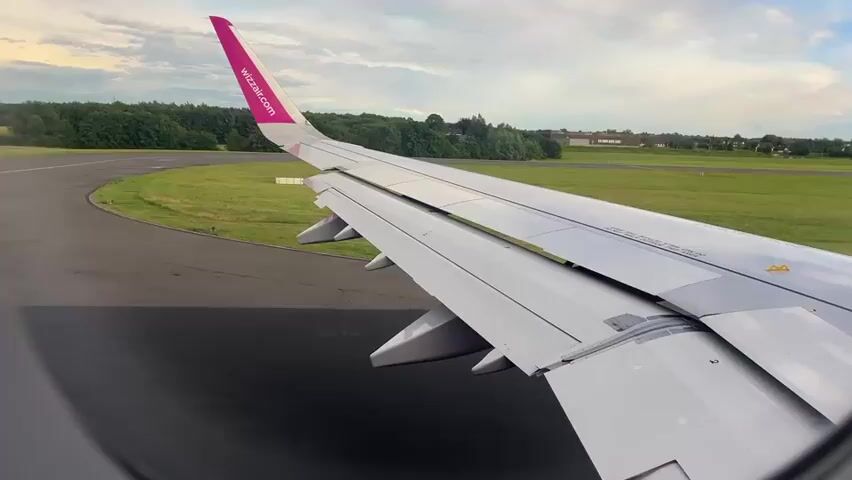 The video provides a view from an airplane window, showcasing the right wing of a Wizz Air aircraft. The plane appears to be on the ground, possibly taxiing or waiting at an airport. The wing is the dominant subject, with its sleek, grey surface and the prominent pink wingtip (sharklet). The text "wizzair.com" is clearly visible on the pink wingtip.

Beyond the wing, the scene reveals parts of the airport environment. A dark grey asphalt runway or taxiway curves into the foreground, leading past a patch of vibrant green grass. In the middle distance, a line of lush green trees forms a natural barrier, beyond which some low-rise buildings are faintly visible under a soft, overcast sky. The sky itself is a mix of light grey and white clouds, suggesting a cloudy but bright daytime, possibly late afternoon or early evening.

The airplane's flaps are partially extended on the trailing edge of the wing, a common configuration during taxiing, takeoff, or landing. There are no people visible in the frame. The overall impression is one of a quiet moment at an airport, with the aircraft preparing for or concluding a flight. The location context provided is Holzwickede, Germany, which implies the footage was captured at Dortmund Airport (DTM).