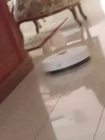 A white robotic vacuum cleaner with a red button is seen moving across a tiled floor in a room. The vacuum is positioned at an angle, with a chair and a wooden table visible in the foreground. Reflections of furniture can be seen on the shiny floor.