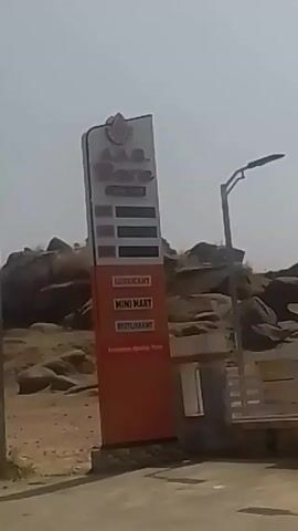 The video depicts a roadside scene in a sparsely populated area. The video shows a gas station with a large sign reading "A.A.G. Bare", which also lists "Lubricant", "Mini Mart", and "Restaurant". A price board is present, though the prices are unreadable. The gas station sits near a rocky outcrop in a dusty, arid environment. Beyond the gas station, there is a road on which a car and motorcycle travel. A sign reading "A.A.G. Bare" is visible along the side of the road. Utility poles line the road. Further down the road, a building under construction can be seen. The sky is bright and hazy, suggesting a sunny day.