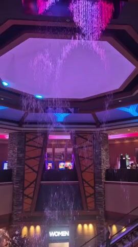 The video shows an indoor waterfall feature. The water falls from an octagonal ceiling piece illuminated with purple, blue, and yellow lights. The water cascades down towards a lower level, with the word "WOMEN" visible on a sign. The surrounding area features stone and wooden architectural details. Slot machines are visible in the background. An escalator is also present.