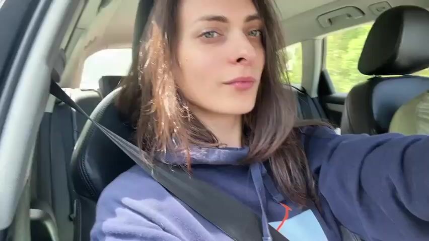 A woman is sitting in the driver's seat of a car, looking at the camera. She has long, dark brown hair and is wearing a dark blue hoodie. A black seatbelt is visible across her chest. The interior of the car is visible, including leather seats and the dashboard. The scene appears to be outdoors, with greenery visible through the car windows. The woman smiles and closes her eyes briefly before opening them again. The video is captured from a selfie perspective. Another person's head and shoulder, wearing a green shirt and a baseball cap, are visible in the passenger seat.