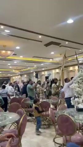 The scene is an interior shot of a brightly lit reception hall. Multiple people stand around tables with pink cushioned chairs. A small child wearing a jacket and jeans is walking around near the tables. Some of the standing adults are taking photos and videos. There are men in traditional clothing holding flags or staffs near a large decorative white floral display. There are ornate lighting fixtures and ceiling decorations throughout the room.