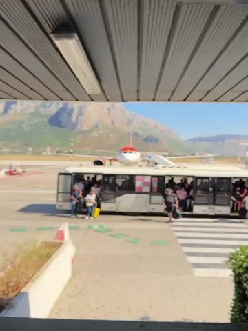 The video shows a bus parked at an airport terminal, with passengers disembarking onto the pavement.  In the background, an orange and white passenger jet sits on the tarmac with stairs attached to the front entrance. Behind the jet, a large mountain dominates the horizon under a clear blue sky. The camera view is from within a covered structure, partially blocking the upper frame of the image.  People are seen walking across the crosswalk after exiting the bus. Other vehicles are visible in the background, along with airport runway markings and signaling equipment.