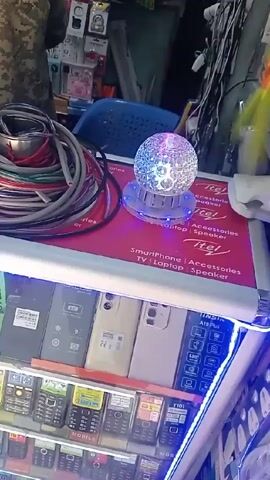The video shows a display case with several rows of mobile phones. There are a few smartphones on top of the display, along with a bundle of wires in a metal container and a disco ball-like lamp on a red surface. A person in camouflage clothing is standing in front of the display case. The display advertises Itel smartphones, laptops, TVs, and speakers. There is text on the phone screens, indicating their prices. The lamp changes colors. The phones on display are various models, including some feature phones with numeric keypads.