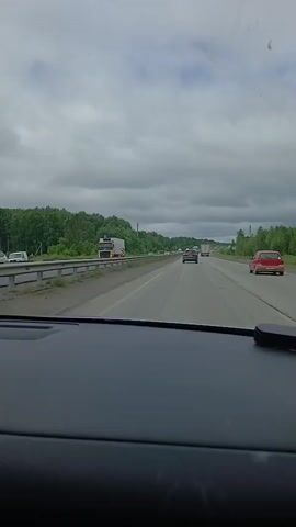 The video is shot from inside a car, looking out through the windshield at a highway. The sky is overcast with gray clouds. On the left side of the highway, there is a guardrail and then a line of trees. On the highway, there are several vehicles, including cars and trucks, traveling in the same direction. A white truck with a yellow and orange striped trailer is visible on the left side of the highway. A red car is in the right lane. The road appears to be in good condition. The overall scene suggests a typical day of travel on a highway.