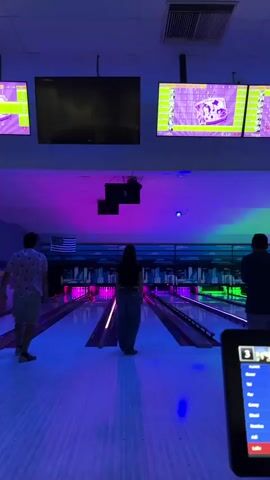 The video shows a group of people at a bowling alley. The lighting is dim and colorful, with purple and blue lights illuminating the lanes. There are several large screens displaying scores and graphics. In the foreground, a person is holding a laptop, partially obscuring the view of the bowling lanes. In the middle ground, a woman is standing on a bowling lane, facing away from the camera. She is wearing baggy jeans and a black crop top. To her left, a man in a patterned shirt is walking away from the camera. To her right, a man in a dark blue shirt is also walking away. In the background, several bowling lanes are visible, with pins at the end of each lane. The overall atmosphere appears to be casual and fun.
