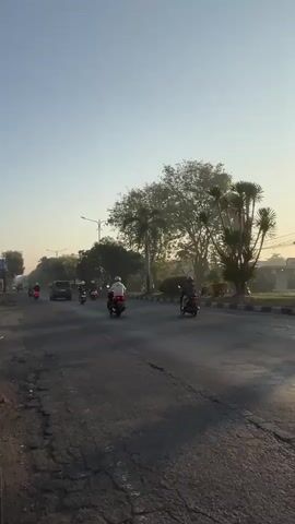 The video shows a street scene with several motorcycles and a truck. The sky is clear and the sun appears to be low in the sky, casting long shadows. On the right side of the road, there are trees and some buildings in the distance. The road itself appears to be asphalt and is in fair condition with some cracks. Several motorcycles are visible, some carrying one rider and others carrying two. One motorcycle in the foreground has a large, rectangular basket strapped to the back, which appears to be carrying goods. A truck is also visible in the distance. The overall impression is of a busy road during what appears to be either early morning or late afternoon.