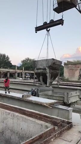 In Khasavyurt, Russia, a crane is lifting a large, rectangular concrete container. A man in a red shirt and dark pants stands to the left, observing the operation. Another person is visible near the concrete structure beneath the hanging container. The scene appears to be an outdoor construction or industrial site, with concrete structures and metal elements suggestive of manufacturing or repair work. The sky indicates it is either early morning or late evening, with a soft, diffused light and hints of pink and orange clouds. The overall atmosphere seems calm and functional, focused on the task at hand. There is no visible text in the media.