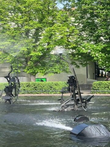 The video showcases a vibrant outdoor scene centered around a large, kinetic fountain in a public square or park. The fountain is composed of multiple black metal sculptures, appearing to be abstract mechanical contraptions made from industrial or recycled parts like large wheels, levers, pipes, and scoops.

The sculptures are actively moving, and water is continuously spraying and splashing from various points, creating a dynamic and interactive display. Lush green trees, some with light-colored leaves, form a verdant backdrop, with several buildings visible beyond them. The weather is sunny, indicating daytime.

In the background, daily life unfolds: a person cycles past on the left side of the frame, and another individual is seated on a low wall or bench. Decorative hedges and a flowering wisteria vine (purple blossoms) add to the pleasant atmosphere. Partial text visible on a building sign in the background reads "Restaurant...halle."