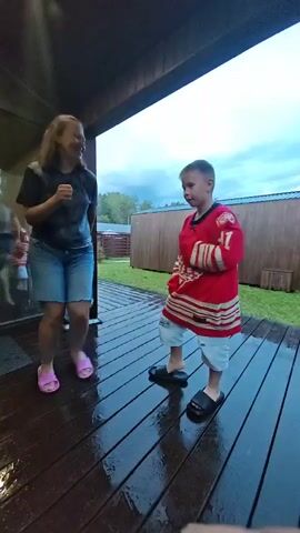 A person with long hair, wearing a dark t-shirt and denim shorts, is dancing and gesturing with their hands on a wet wooden deck under a covered patio. They are wearing pink flip-flops. A young boy, wearing a red hockey jersey with the number 91 and light blue shorts, is standing facing the person and appears to be observing them. He is wearing black slides. The deck is wet, suggesting it has recently rained or is raining. In the background, there is a wooden fence and some greenery. A glass door or window is visible to the left, with reflections of people moving. The sky appears overcast.