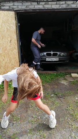 The video is filmed vertically. In the foreground, a person with long blond hair is smiling and looking at the camera. They are wearing a white t-shirt and black shorts with red trim. Behind them, a person is sitting in the driver's seat of a dark-colored car parked inside a garage. The garage has a concrete floor and brick walls. The person in the car is wearing a gray t-shirt and dark pants. They appear to be working on something inside the car. The garage door is open, and the background is an outdoor area with dirt and grass.