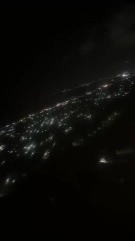 The video shows a nighttime aerial view of a sprawling city illuminated by countless lights. The lights appear as bright white and yellow points scattered across a dark landscape, suggesting a densely populated urban area. Some areas of the city seem more brightly lit than others, possibly indicating commercial districts or major thoroughfares. The camera angle suggests the viewpoint is from an airplane or a similar elevated position. The overall impression is one of a vast, active metropolis seen from above in the dark.