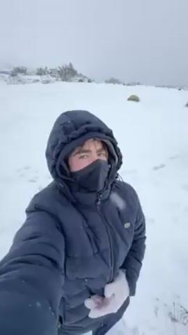 A young person wearing a dark blue puffy jacket, a black mask, and gloves stands in a snowy landscape. The person then turns and walks away, carrying a red object, possibly a plank. The camera pans across a field of snow with light vegetation, rocks, a fence, and trees in the distance. The sky is a uniform light gray, suggesting overcast conditions.